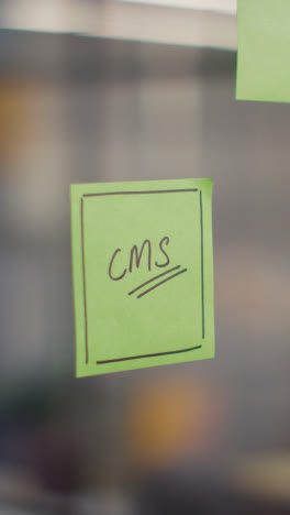 Vertical-Video-Close-Up-Of-Woman-Putting-Sticky-Note-With-CMS-Written-On-It-Onto-Transparent-Screen-In-Office